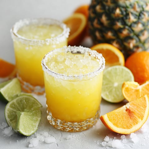 Two glasses of Cinco De Drinko sit on a table with a pineapple and oranges.